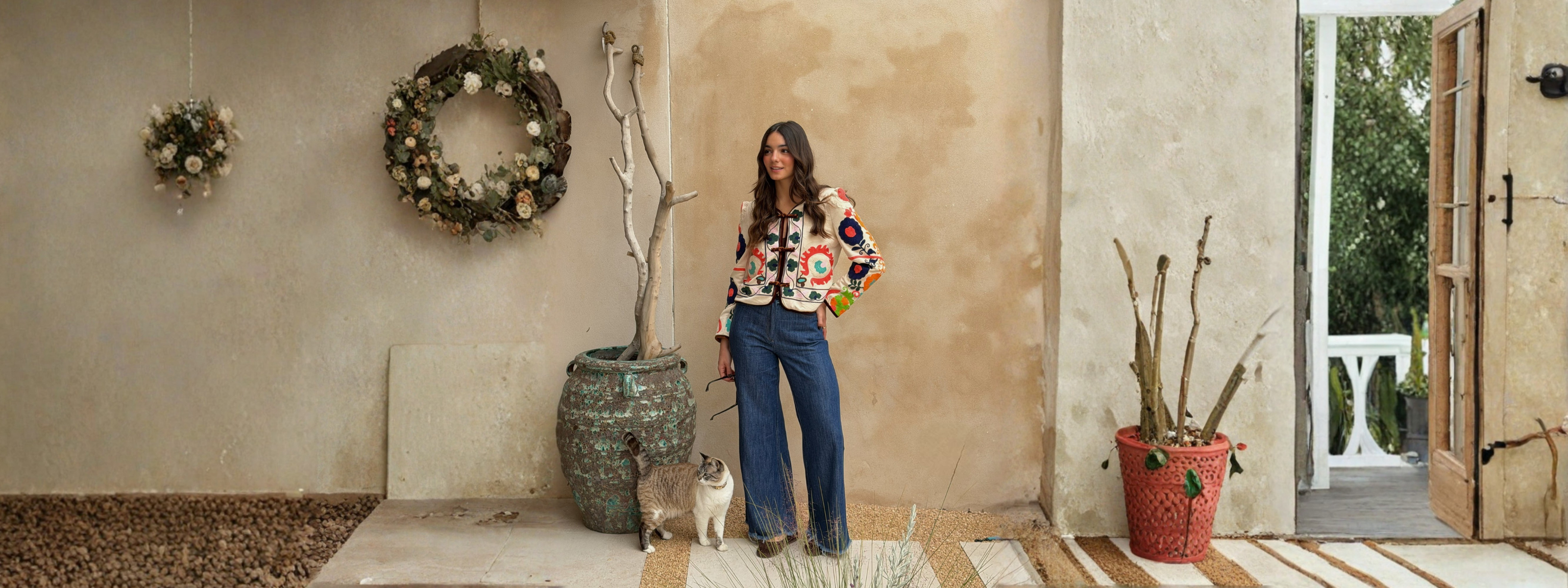 Mujer con abrigo colorido y pantalones azules, posando en un entorno acogedor con gato y decoración de temporada.