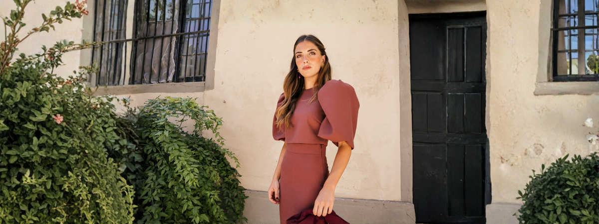 Conjunto de Invitada en Mariquita Trasquilá, mujer con conjunto de invitada en tono teja, con top de mangas abullonadas y falda de cintura alta, posando junto a una fachada rodeada de vegetación.