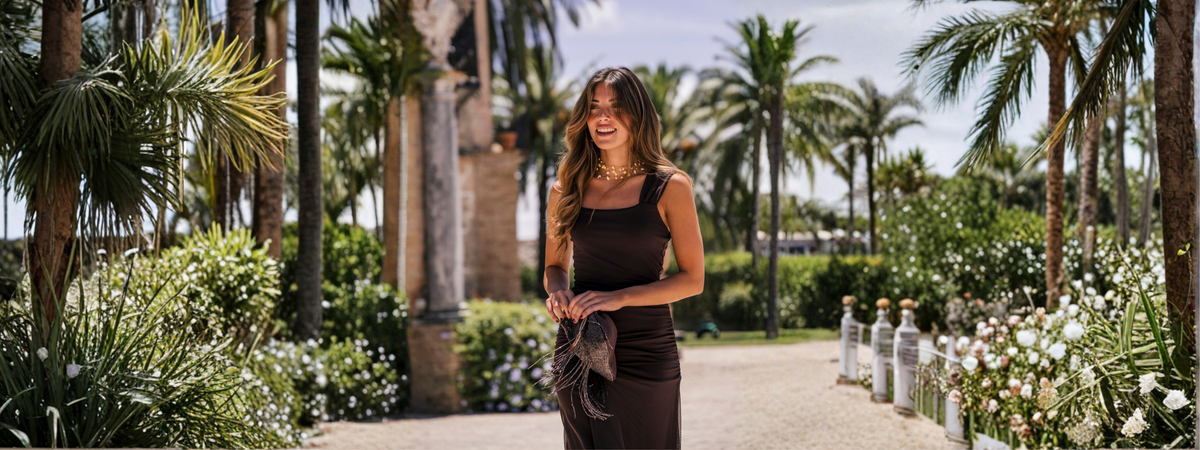 Mujer con vestido largo marrón caminando por un jardín soleado rodeado de palmeras, flores y vegetación.