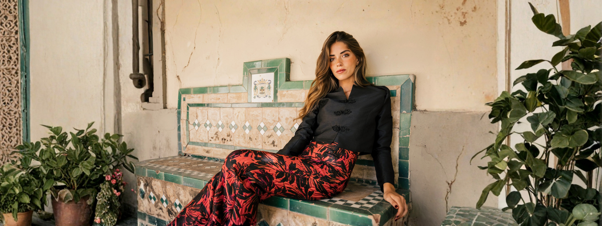 Mujer con chaqueta negra y falda larga de estampado floral rojo y negro, sentada en un banco de azulejos rodeado de vegetación.