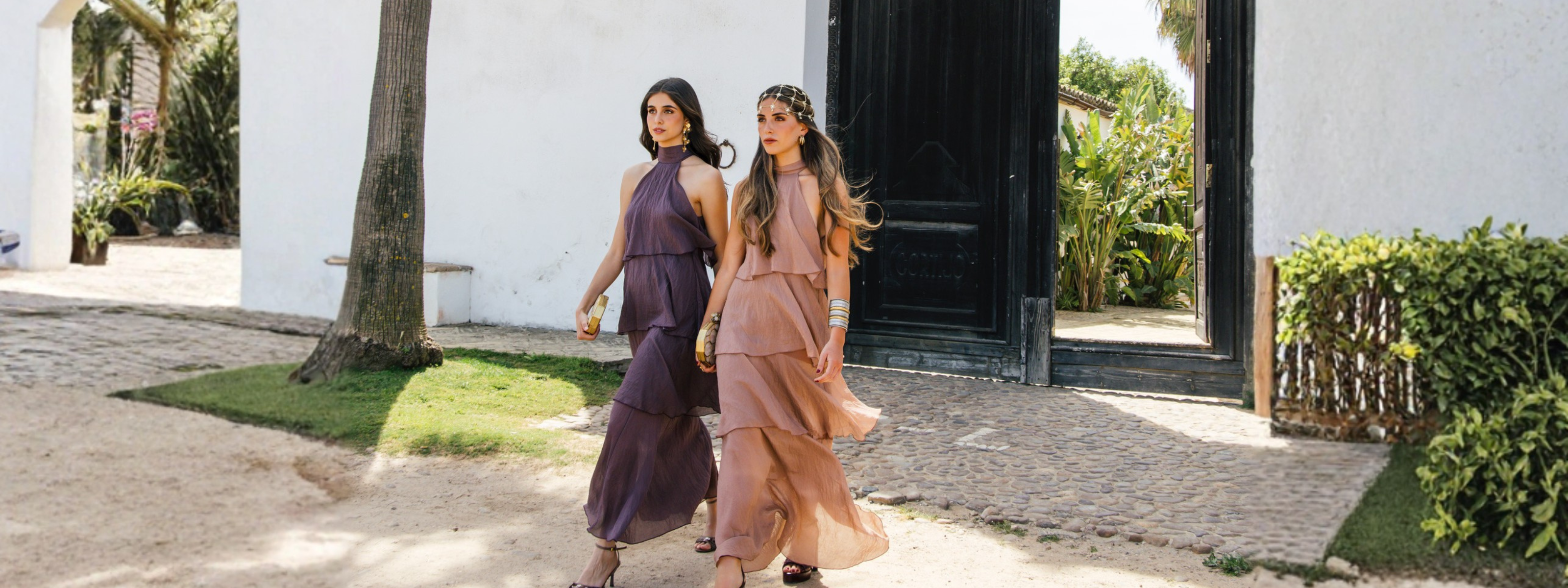 Dos invitadas de verano caminando por un patio exterior con diseños fluidos de volantes: una en tono morado berenjena y otra en rosa empolvado, ambas con sandalias de tacón y complementos llamativos.