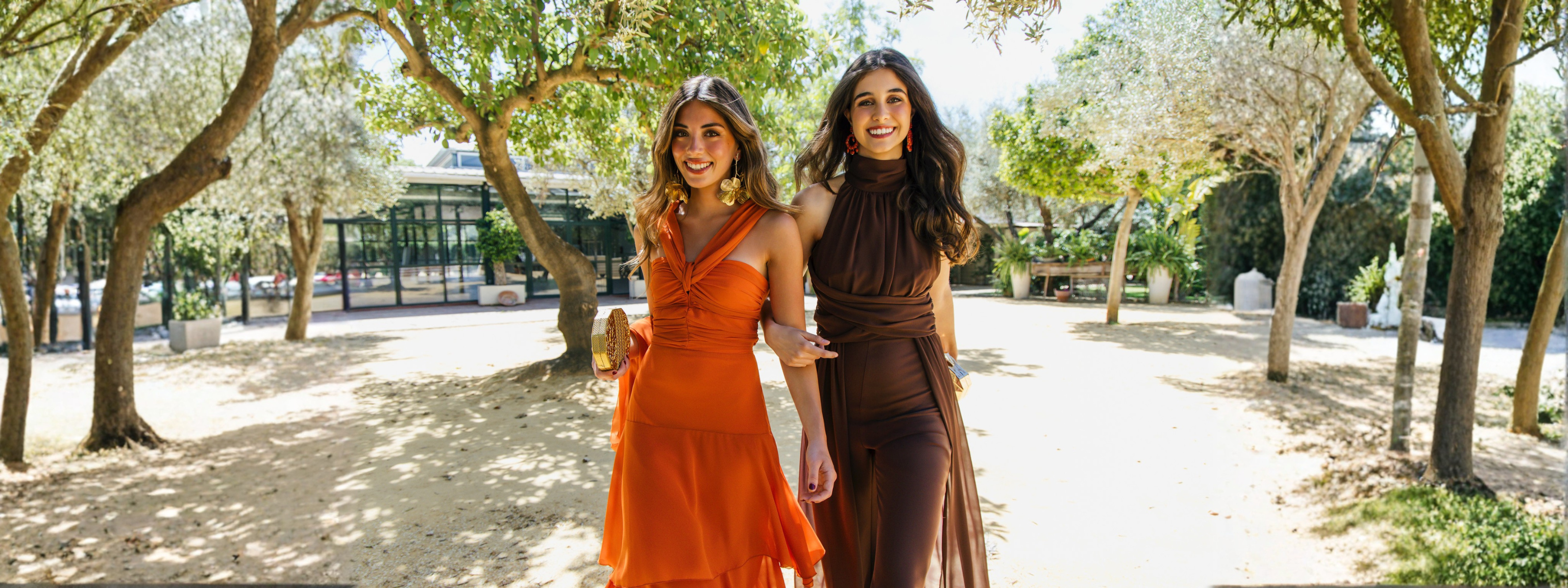 Dos invitadas de bautizo. Una con un vestido naranja de gasa con el cuello cruzado y su amiga con un conjunto dos piezas en marrón también de gasa.