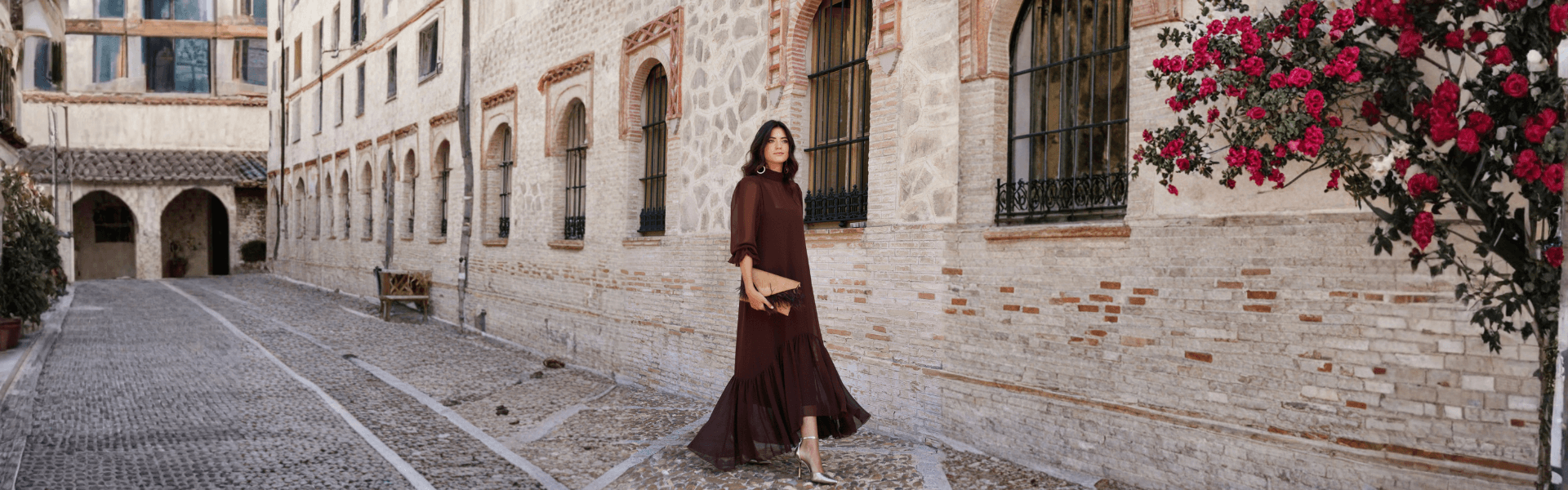 Mujer caminando en la calle con un elegante vestido de fiesta, rodeada de flores en un entorno arquitectónico.