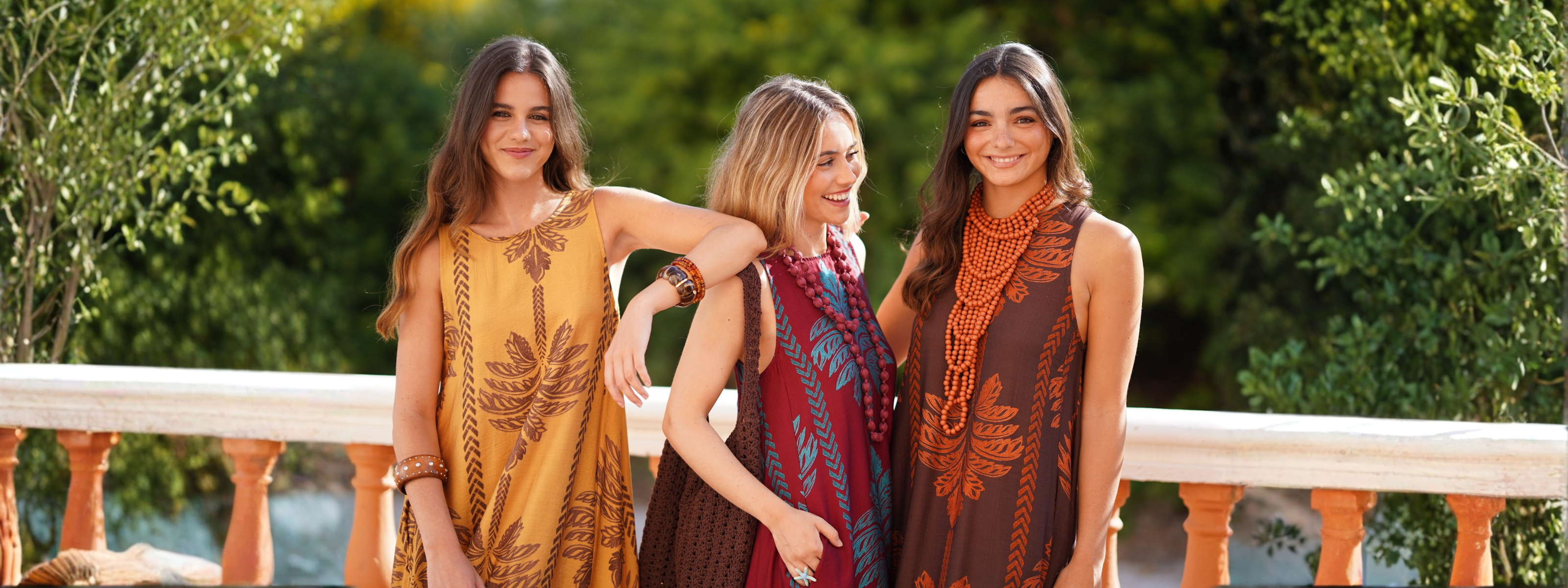 Dos mujeres caminan por un jardín junto al mar con vestidos largos estampados de Mariquita Trasquilá, rodeadas de palmeras y vegetación en un ambiente fresco y veraniego.